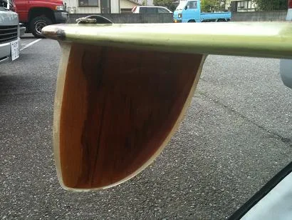 Wide wooden D fin under the tail of a green longboard in a parking lot